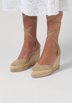 Vrouw met beige suède sleehak espadrilles met enkelbandjes en een witte gebreide rokszoom zichtbaar boven de enkels.