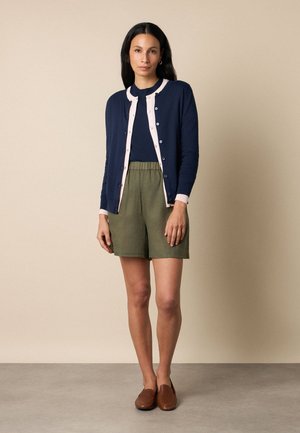 Frau steht, trägt eine marineblaue und hellrosa Cardigan mit Knöpfen, ein marineblaues Oberteil, olivgrüne Shorts und braune Loafers vor beigefarbenem Hintergrund.