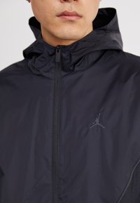 Črna kapuca jakna iz lahkega materiala, ki ima poln zadrgo in diskreten Air Jordan logo na prsih.