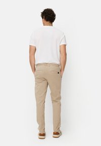 Beige Baumwollhosen mit geradem Schnitt, zwei Gesäßtaschen und Bindebändern an den Knöcheln, kombiniert mit einem schlichten weißen T-Shirt mit kurzen Ärmeln.