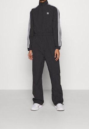 Osoba ubrana w czarny dres Adidas z białymi paskami na rękawach, stojąca na szarej podłodze na tle białej ściany.