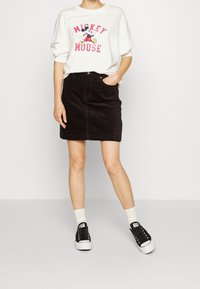 Valkoinen huppari, jossa on punainen "Mickey Mouse" -grafiikka, yhdistetty mustiin farkkuhameeseen ja mustiin lenkkareihin. Malli seisoo kädet taskuissa.