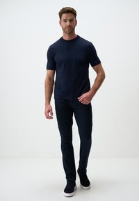 Granatowa koszulka z krótkim rękawem z gładkiego materiału w zestawieniu z ciemnogranatowymi spodniami. Model w ciemnych trampkach, stojący na jednolitym tle.