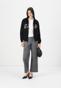 Black zip-up hoodie with floral “GAP” lettering, white fitted t-shirt, gray wide-leg cargo pants, and black pointed-toe heels. Black handbag included.