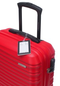 Red polycarbonate suitcase with textured horizontal lines, black handle, and a tag for personal information attached to the side.