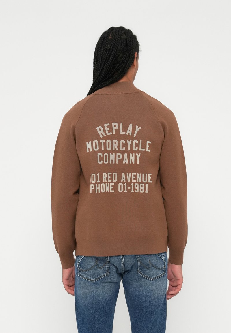 Brun strikket genser med ribbestrikket krage, med hvit tekst på baksiden: "REPLAY MOTORCYCLE COMPANY 01 RED AVENUE TELEFON 01-1981."