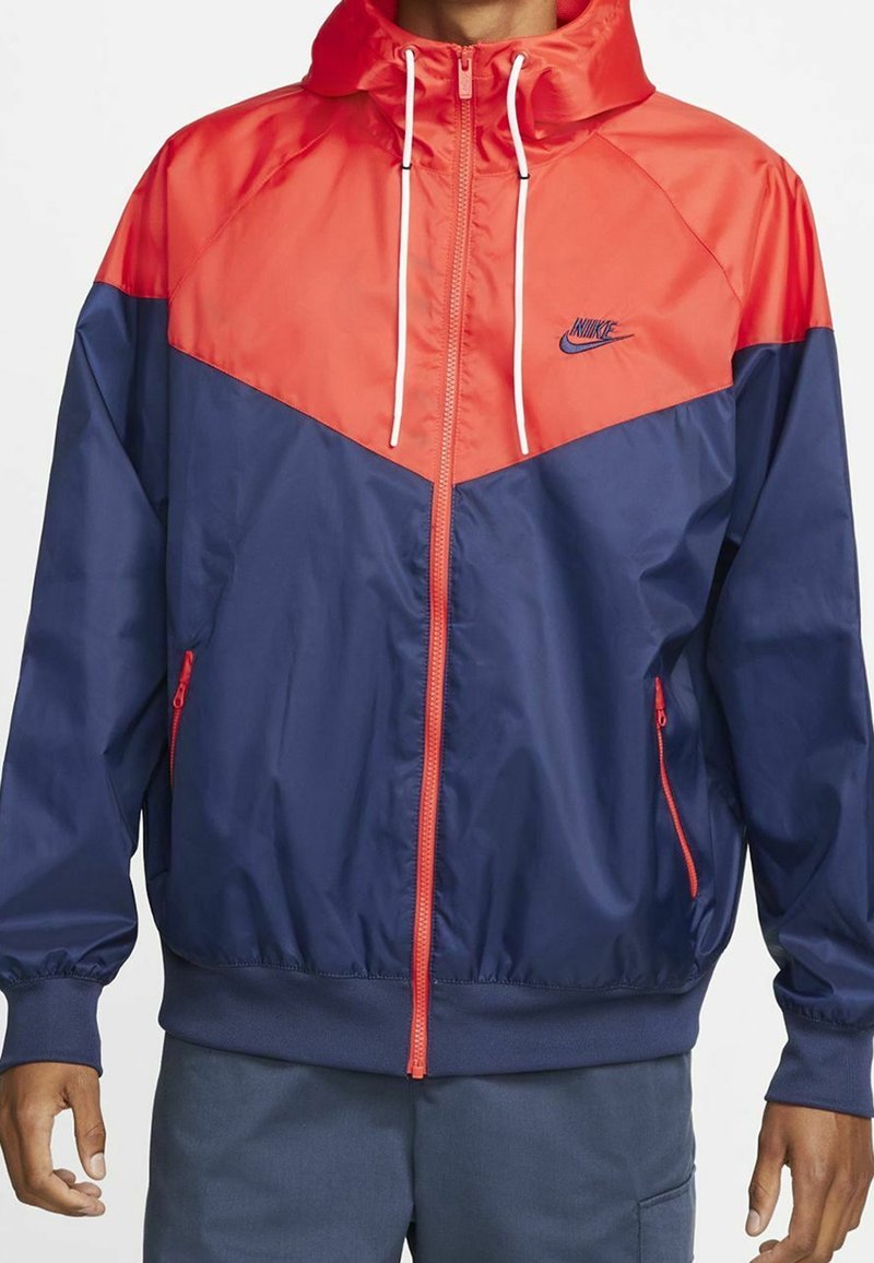 Nike windbreaker jacket in red and navy. Features a zippered front, adjustable hood, elastic cuffs, and two side pockets. Smooth, lightweight material.