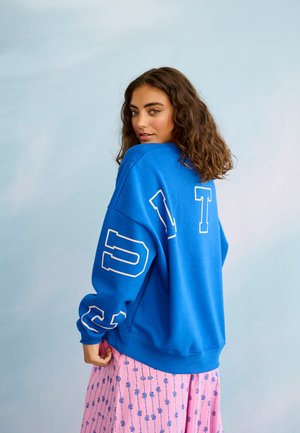 Joven con cabello rizado que lleva una sudadera azul grande con letras blancas y una falda rosa con estampado, mirando por encima del hombro.