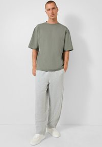 Junger Mann steht in einem übergroßen olivgrünen T-Shirt, hellgrauen Jogginghosen und weißen Turnschuhen vor einem schlichten weißen Hintergrund.