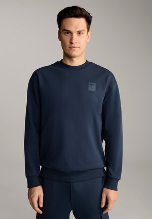 Granatowy sweter z okrągłym dekoltem, z prążkowanymi mankietami i dolnym wykończeniem, z małym wytłoczonym logo na piersi. Miękki, teksturowany materiał.