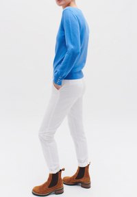 Blauwe trui met een ronde halslijn, aansluitende manchetten met knopen, gecombineerd met witte broek. Model draagt bruine suède enkellaarsjes.