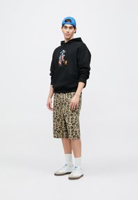 Μαύρο φούτερ με κουκούλα με γραφικό σχέδιο, σορτς με ταμπά λεοπάρδαλη εκτύπωση και λευκά αθλητικά παπούτσια με γκρι λεπτομέρειες. Μια μπλε καπελάκι του μπέιζμπολ ολοκληρώνει το ντύσιμο.