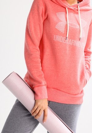 Osoba ubrana w koralową bluzę z kapturem Under Armour i szare spodnie, trzymająca w jednej ręce zwiniętą jasnoróżową matę do jogi.