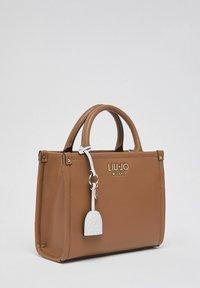 Sac à main en cuir marron avec deux poignées, détails du logo et un porte-clés blanc en accent. Forme rectangulaire avec finition texturée.