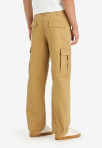 Beige cargo pants with a relaxed fit, featuring side pockets and back flaps. Made of durable fabric, ideal for casual wear.