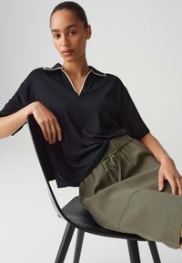 Woman with pulled-back hair sits on black chair wearing black short-sleeve top with white trim and olive green drawstring pants.