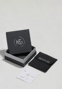 Cutie de bijuterii neagră și argintie cu un logo pe capac, însoțită de un săculeț negru și o etichetă albă cu text și detalii aurii.