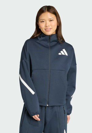 Mornariće plavi dukserica na zip, izrađena od mekanog materijala. Ima bijele pruge na rukavima i Adidas logo na grudima. Opustív fit, sa kapuljačom.