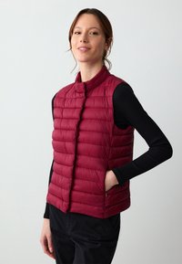 Gilet imbottito borgogna con colletto alto, dotato di sezioni trapuntate e tasche laterali. Realizzato in tessuto leggero, progettato per essere indossato a strati.