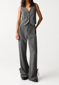 Gilet gris sur mesure avec col en V et trois boutons, associé à un pantalon taille haute à jambes larges avec liens ajustables aux chevilles.