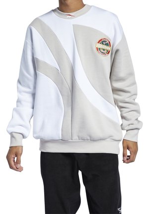 Henkilö pukeutunut valkoiseen ja vaaleanharmaaseen Reebok-collegepaitaan, jossa on abstrakteja kaarevia kuvioita ja pyöreä Reebok Classic -logo rinnassa.