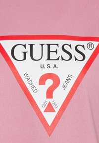 T-shirt en coton rose avec un grand logo triangulaire blanc et rouge portant le texte "GUESS" et des icônes "WASHED" et "JEANS" en texte noir gras.