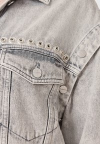 Giacca di jeans grigio chiaro con cuciture nere, caratterizzata da occhielli argentati lungo la spalla e bottoni metallici marchiati. Tessuto testurizzato.