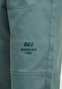Zelené látkové kalhoty s měkkou texturou, které mají potištěný nápis "DEF WORKATION MODE F/W23" na stehně, se zesílenými švy.