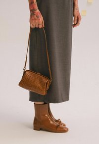 Bolso de mano marrón con estampado de cocodrilo y una correa larga, combinado con botas de tobillo brillantes marrones con un detalle de lazo, usado con un vestido maxi gris.