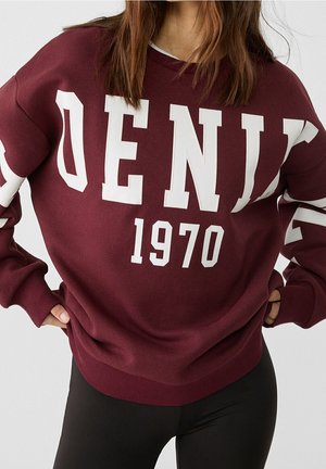 Kobieta w bordowej bluzie z dużymi białymi literami układającymi się w słowo "DENIM" i liczbą "1970", w połączeniu z czarnymi legginsami.
