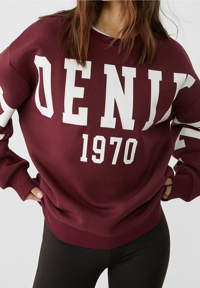Femme portant un sweat-shirt bordeaux avec de grandes lettres blanches formant "DENIM" et le numéro "1970", associé à des leggings noirs.