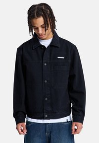 Νέος άνδρας με πλεγμένα κοτσίδες, φοράει μαύρο σακάκι με κουμπιά πάνω από λευκό πουκάμισο και μπλε τζιν, κοιτάζοντας προς τα κάτω.