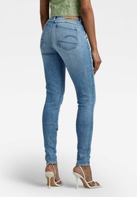 Mujer vistiendo jeans ajustados de color azul claro y sandalias de tacón blanco, de pie con su espalda frente a la cámara.