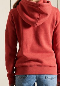 Sudadera con capucha roja hecha de una tela suave, con un cuello redondeado y puños acanalados, combinada con jeans de mezclilla azules.