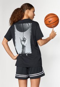 Μαύρο κοντομάνικο μπλουζάκι με γραφικό σε γκρι κλίμακα που απεικονίζει ένα χέρι που κρατάει μια μπάλα μπάσκετ στην πλάτη. Συνδυάζεται με μαύρα αθλητικά σορτς.