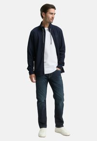 Marineblauwe jas met rits en hoge kraag, gedragen over een wit T-shirt, gecombineerd met donkere jeans en witte sneakers. Soepele stoftextuur.