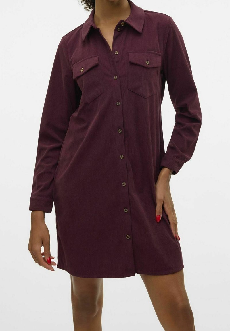 Femme portant une robe-chemise en velours côtelé violet foncé à manches longues, boutonnée, avec deux poches poitrine et du vernis à ongles rouge.