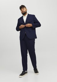 Marineblauer Anzug mit tailliertem Schnitt, einreihigem Sakko, zwei Taschen und passender Hose. Kombiniert mit einem weißen Hemd und schwarzen Sneakers.