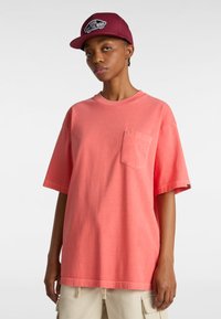 Korallenfarbenes Oversized-T-Shirt aus Baumwolle mit einer Brusttasche und kurzen Ärmeln, kombiniert mit einer kastanienbraunen Kappe mit einem Logo.