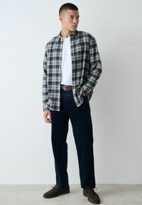 Plaid flannel shirt in black, grey, and white; white t-shirt underneath; dark pants; brown shoes; standing pose against a neutral background.