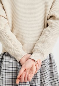 Beige bordás kötött pulóver hosszú, texturált ujjal, világoskék és fehér árnyalatú kockás szoknyával párosítva. A kezek a hát mögött összekulcsolva.