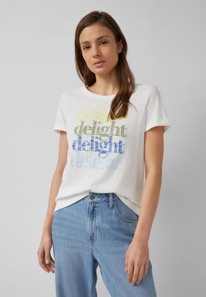 Vrouw met lang bruin haar die een wit T-shirt draagt met meerkleurige "delight" tekst en lichtblauwe spijkerbroek, staande tegen een grijze achtergrond.