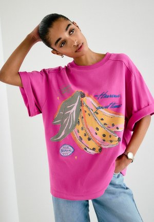Frau mit zurückgebundenem Haar, die ein leuchtend pinkes, übergroßes T-Shirt mit einem bunten Bananenmotiv und blaue Jeans trägt, posiert mit einer Hand am Kopf.