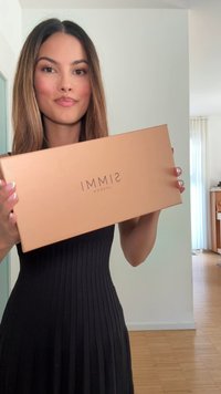 Kobieta z długimi włosami w czarnej sukience trzyma brązowe pudełko po butach SIMMI London w jasnym, nowoczesnym pokoju z drewnianymi podłogami.