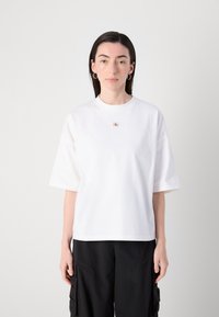 Hvit bomulls t-skjorte med korte ermer, med en liten brodert logo på brystet. Glatt tekstur og avslappet passform.