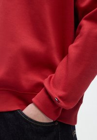 Κόκκινο ριμπ ζακέτα με στρογγυλεμένο λαιμό και μανίκια με μανσέτες. Φέρει ένα μικρό τρίχρωμο λογότυπο στη μανσέτα. Φορεμένη πάνω από σκούρο τζιν παντελόνι.