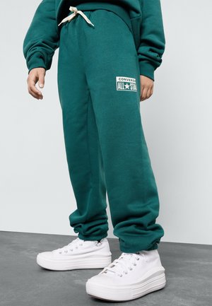 Osoba nosząca turkusowe spodnie dresowe Converse z logo „All Star” i białe wysokie trampki Converse stojąca na szarej podłodze.