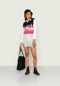 Mulher com um chapéu preto de bucket, um colete sem mangas com padrão de chamas rosa e preto sobre uma camisa branca, saia prateada, botas pretas, segurando uma bolsa tote preta.