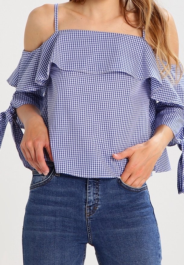 Femme portant un chemisier bleu et blanc à carreaux, épaules dénudées avec volants et manches nouées, associé à un jean en denim bleu.