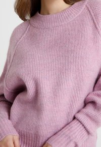 Suéter de punto de color rosa claro con textura acanalada, cuello redondo y mangas largas. Presenta un ajuste relajado y sutiles variaciones en el tono.